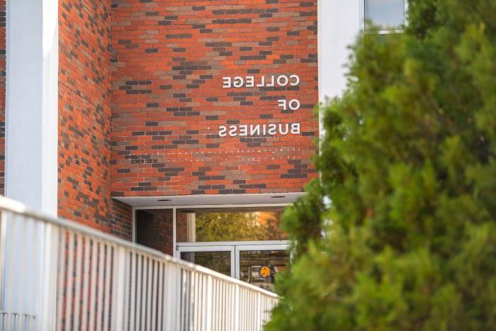 The entrance to a College of Business building, featuring a red and brown brick facade. Large white letters above the door read "College of Business." A forefront tree partially blocking the left side of the image, 在右边可以看到金属栏杆.